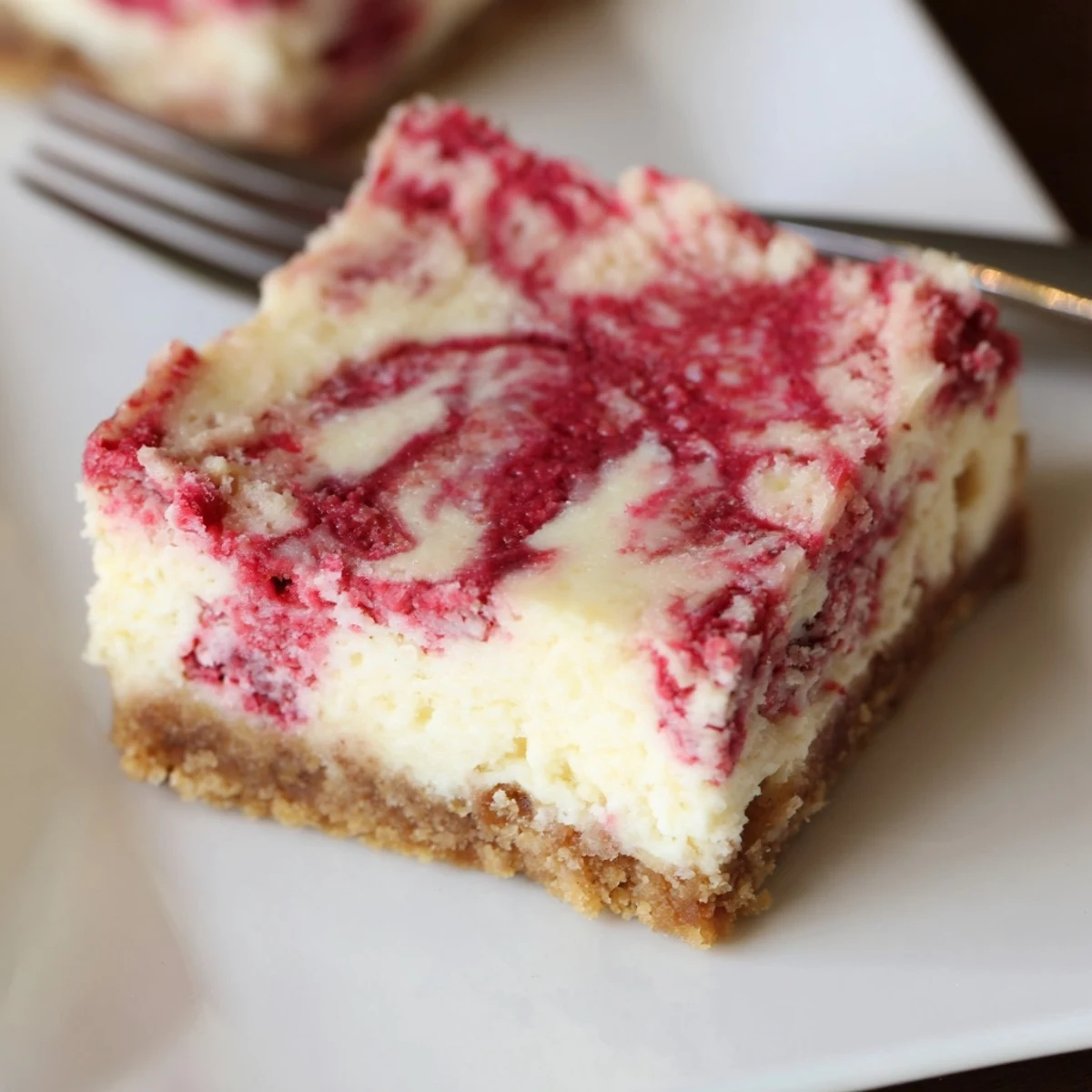 Homemade Raspberry White Chocolate Cheesecake Bars with a creamy filling and graham cracker crust, sliced on a plate.