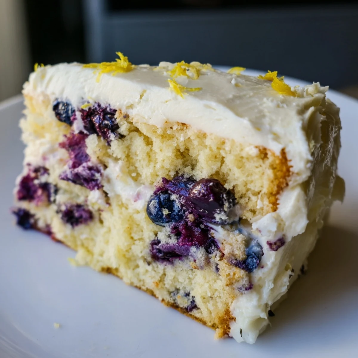 Powdered sugar is being sprinkled over a frosted Lemon Blueberry Cake, highlighting the zesty yellow crumbs and fresh berries on top.