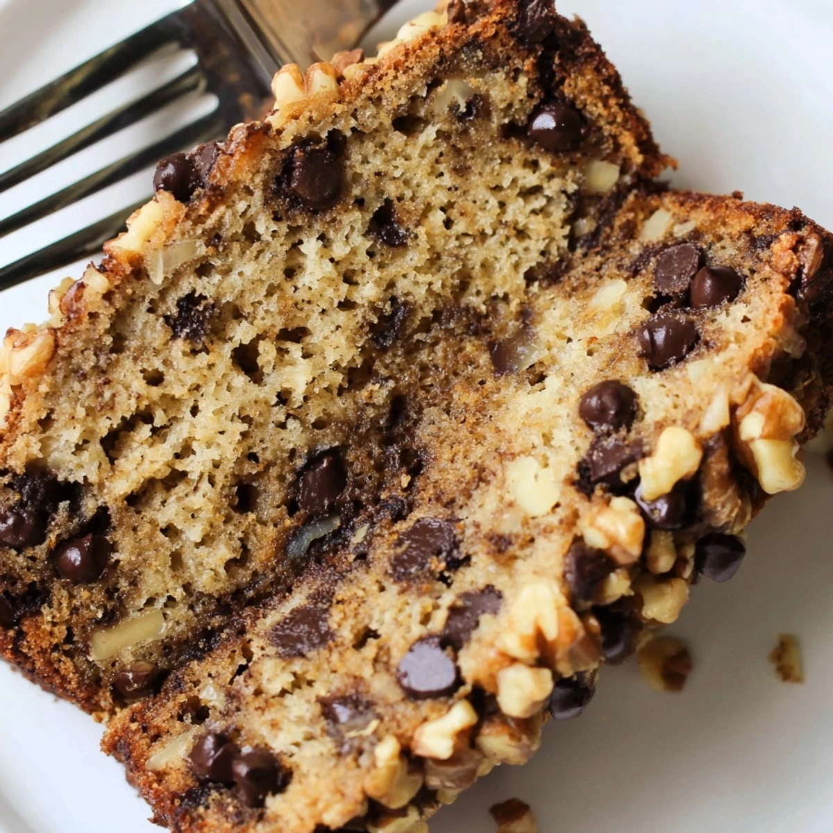 A golden-brown Chocolate Chip Banana Bread with Walnuts loaf sits beside sliced bananas and walnuts on a marble counter.