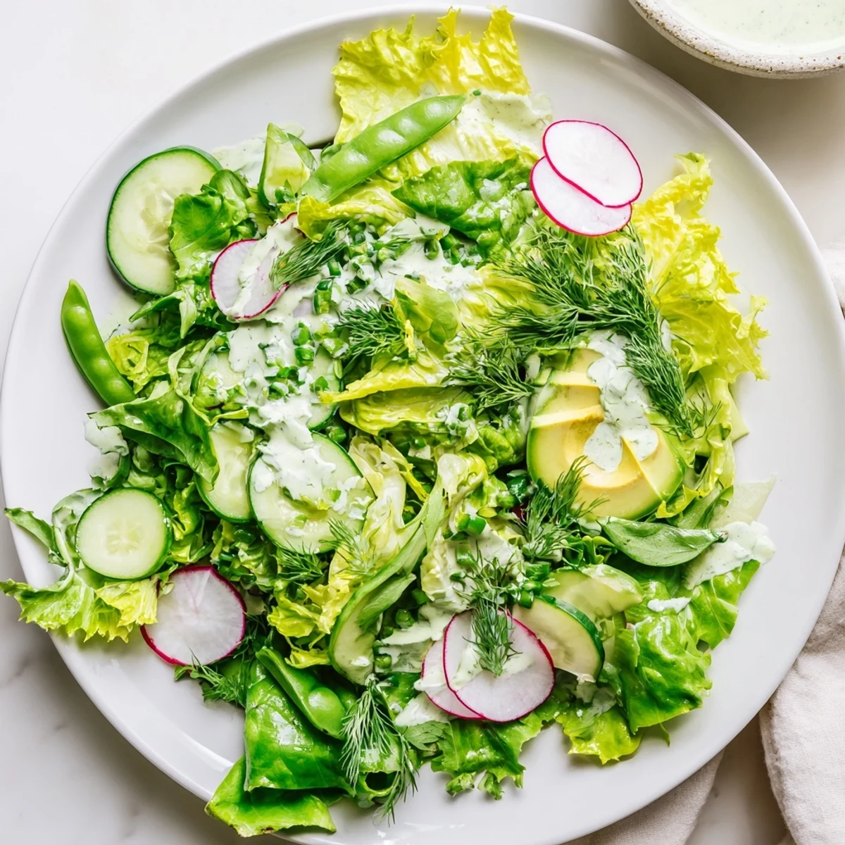 Vibrant bowl of Green Salad with Green Goddess Dressing, featuring snap peas, radishes, and fresh herbs on a marble counter.