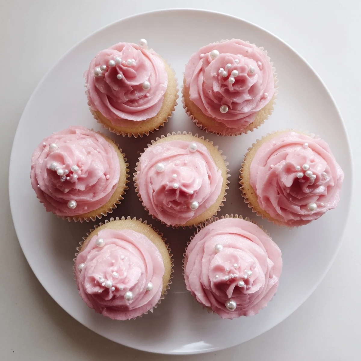A close-up of pink buttercream Girl Baby Shower Cupcakes topped with edible pearls and sprinkles on a pastel stand.