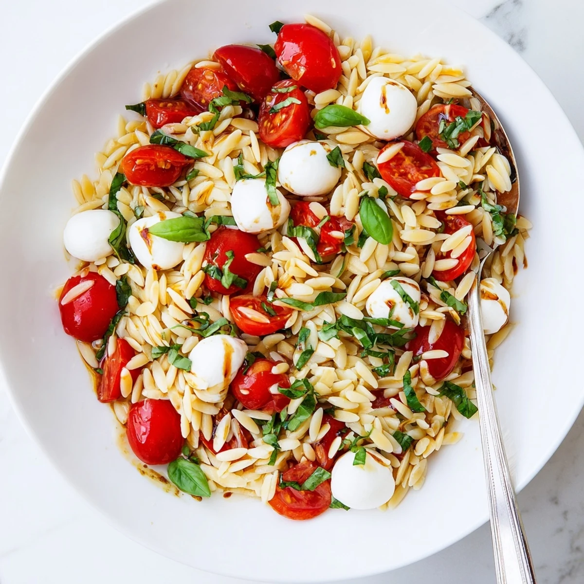 Colorful bowl of Caprese orzo pasta salad dotted with vibrant red tomatoes and white mozzarella balls, drizzled with dark balsamic dressing