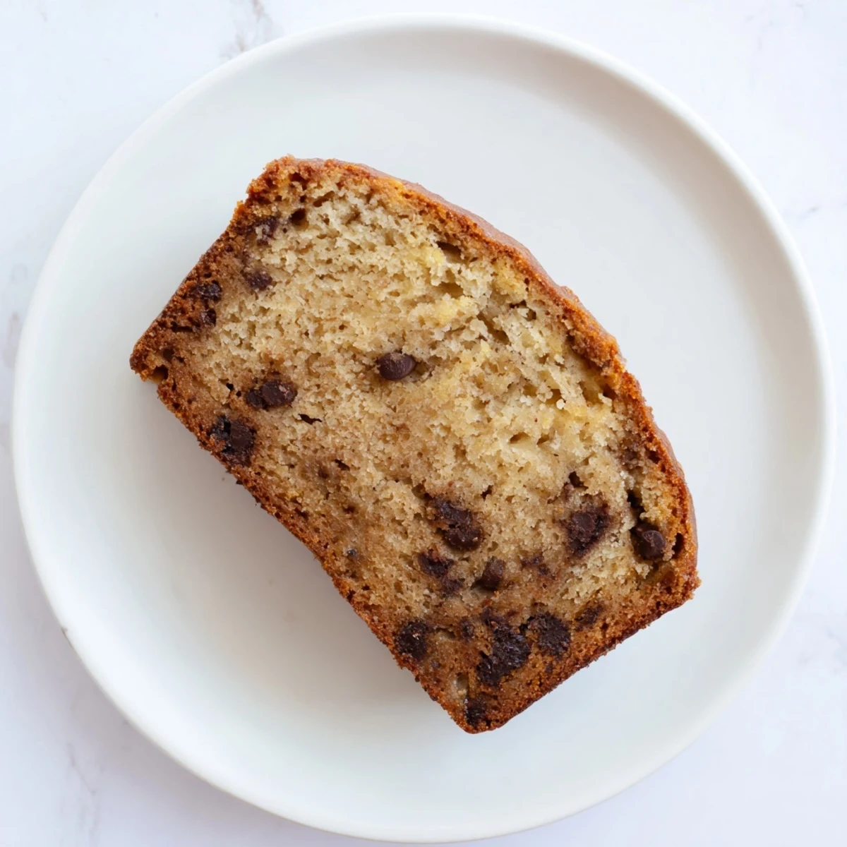 Thick slice of moist chocolate chip banana bread on a white plate with crumbs
